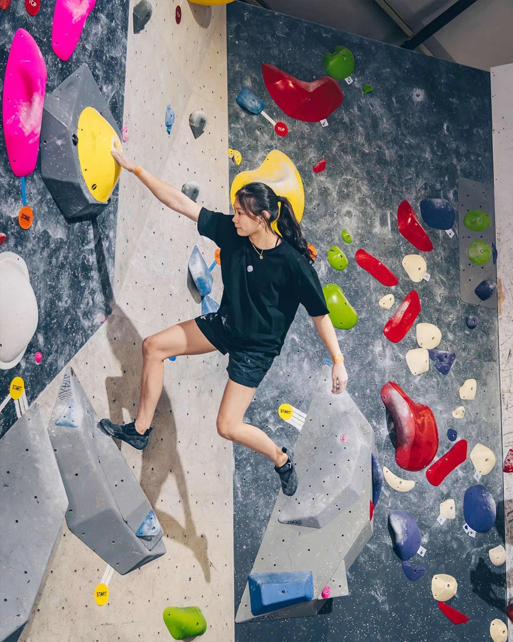 Female climber making a dynamic move on a colourful indoor bouldering wall with START markers visible