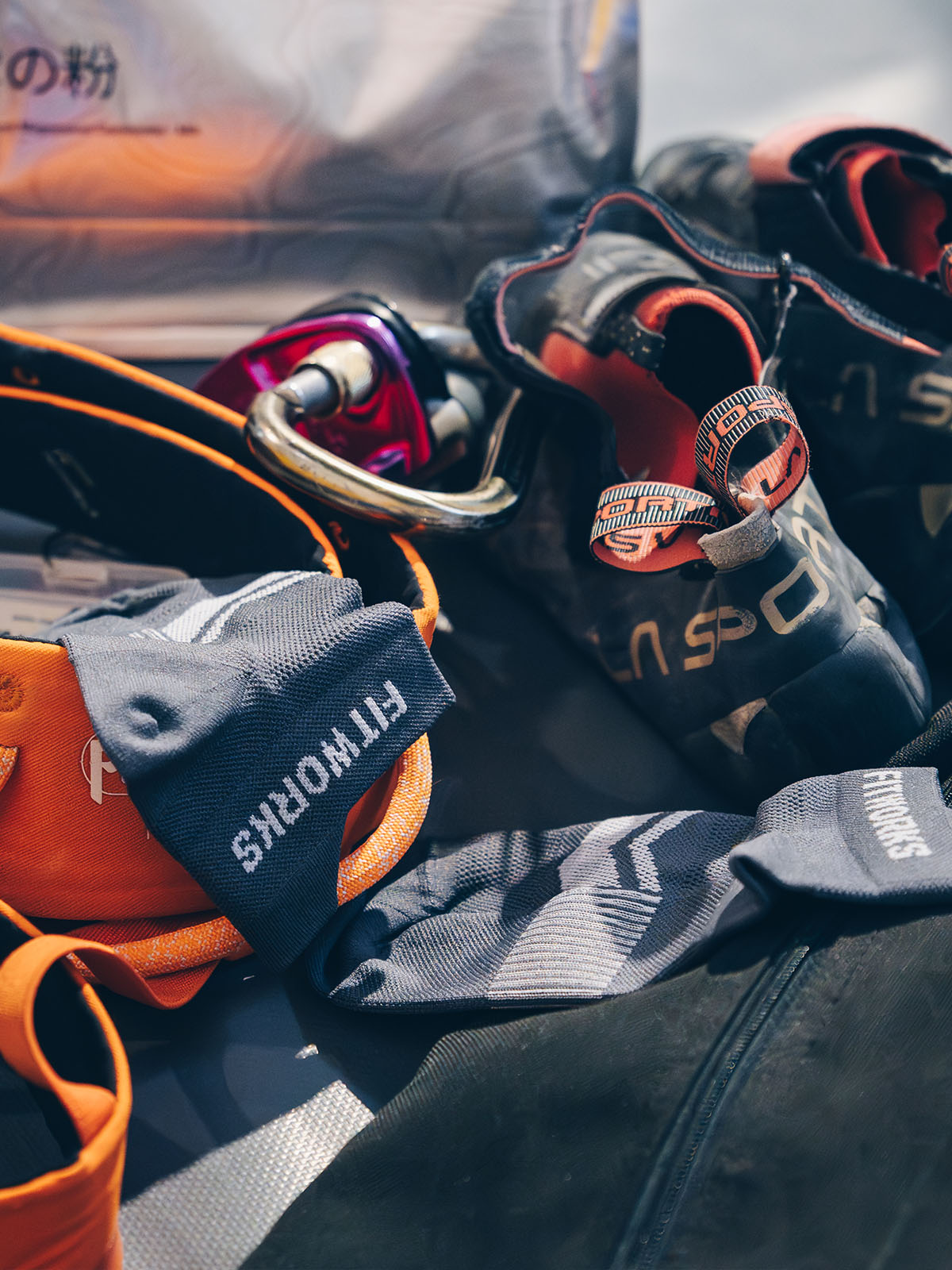 A close-up, angled view of climbing gear including a dark gray climbing shoe with orange lining, a climbing harness, a carabiner, and a gray climbing glove with 'FITWORKS' printed in white. The background shows a soft-focus outdoor scene.