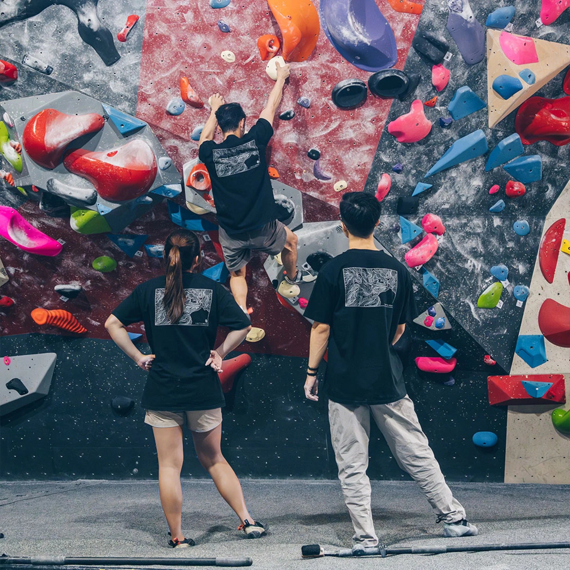 Climber reaching for a hold while two others watch from the mat on a colourful bouldering wall