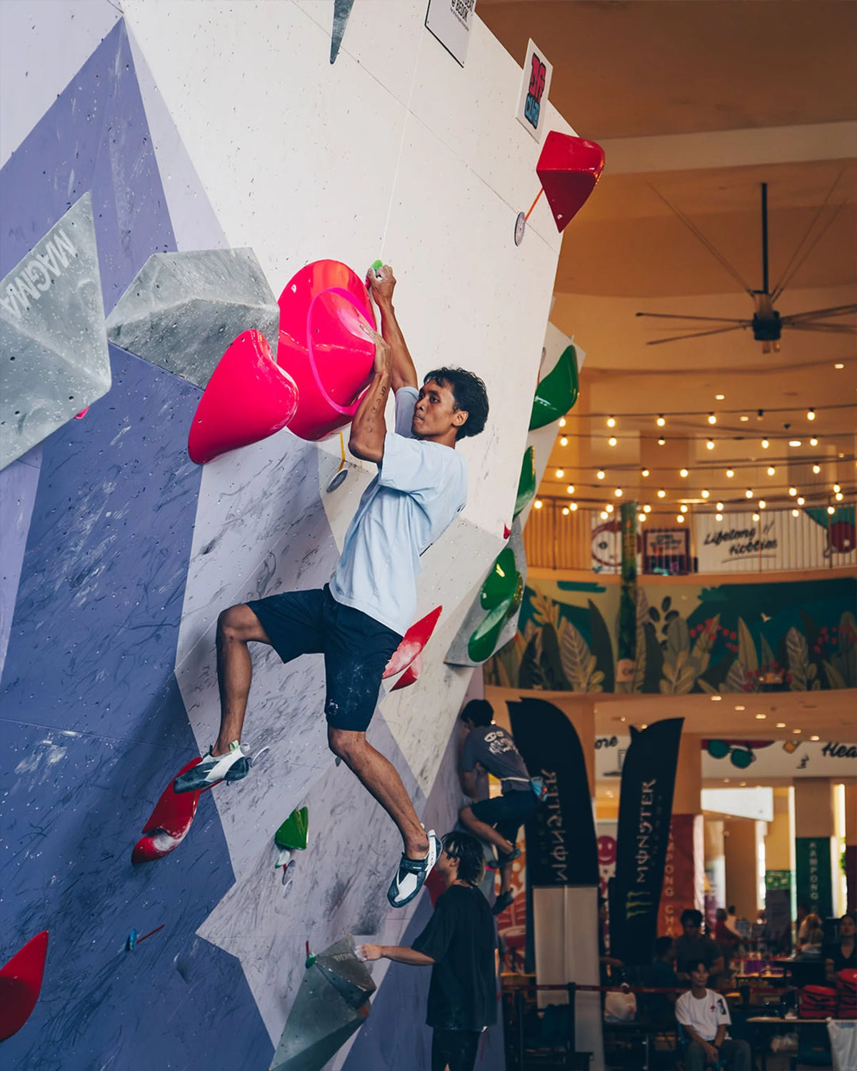 Male climber gripping large pink volumes on a competition bouldering wall set up in a mall atrium