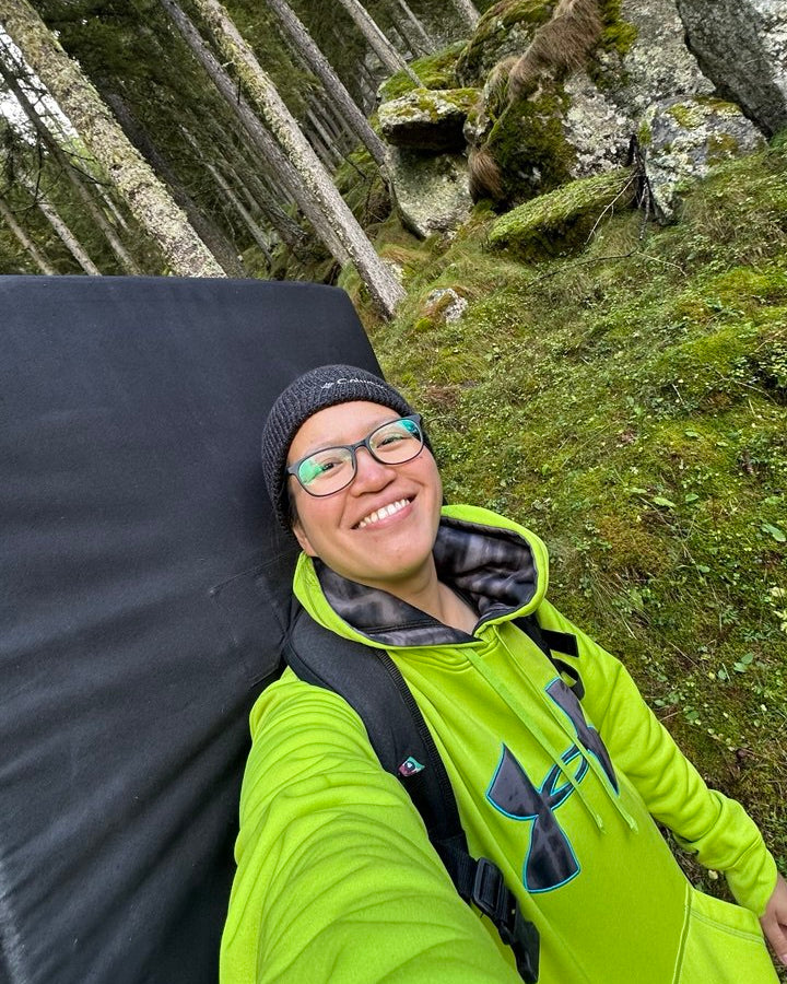 Person in a green jacket with a backpack taking a selfie in a forested area.